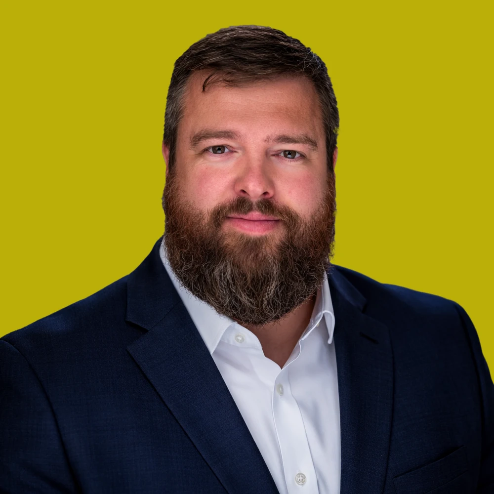 A man with a full beard and short hair, wearing a dark suit jacket and white shirt, poses in front of a plain yellow background.