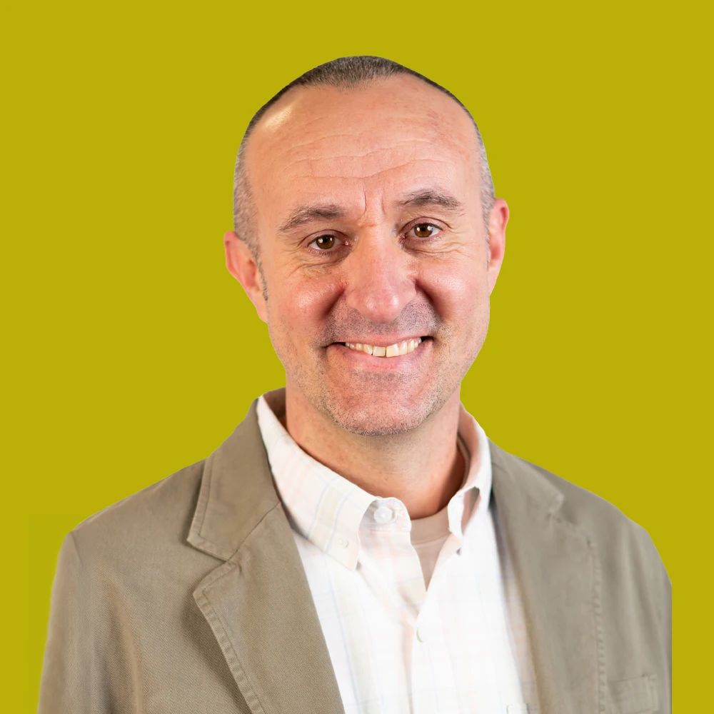 Smiling middle-aged man with short hair, wearing a light-colored blazer and collared shirt, standing in front of a solid yellow background.