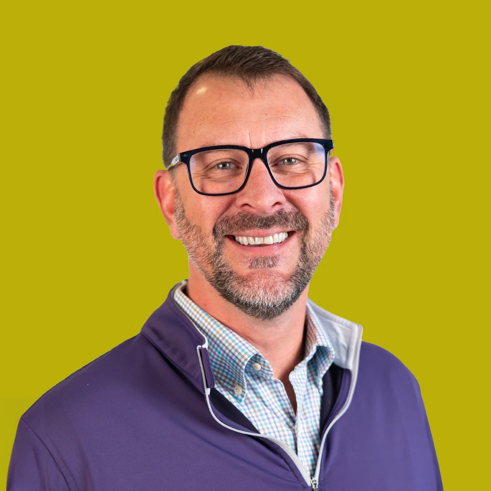 Smiling man with glasses, short hair, and a beard, wearing a purple jacket over a collared shirt, poses in front of a solid yellow-green background.
