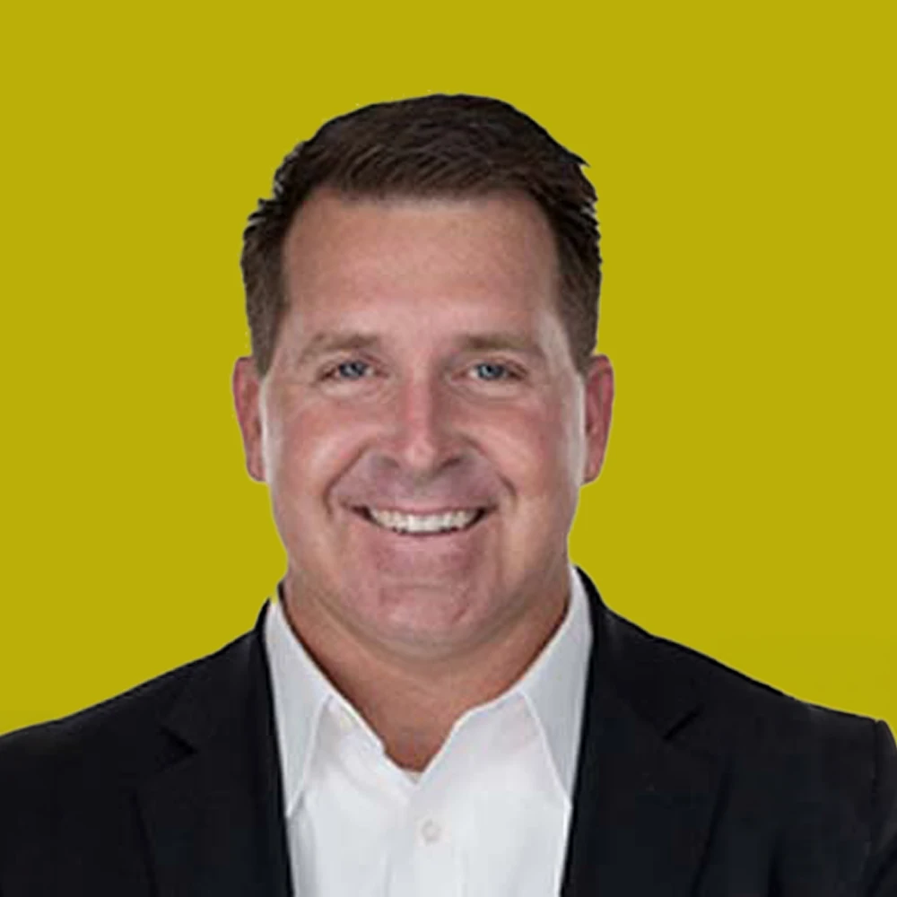 Smiling man wearing a black suit jacket and white shirt, pictured against a plain yellow background.