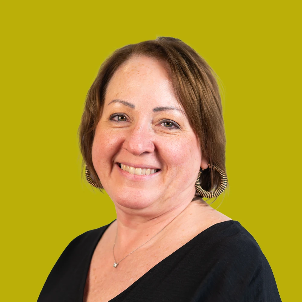 Woman with short brown hair, hoop earrings, and a black top smiles in front of a plain yellow-green background.