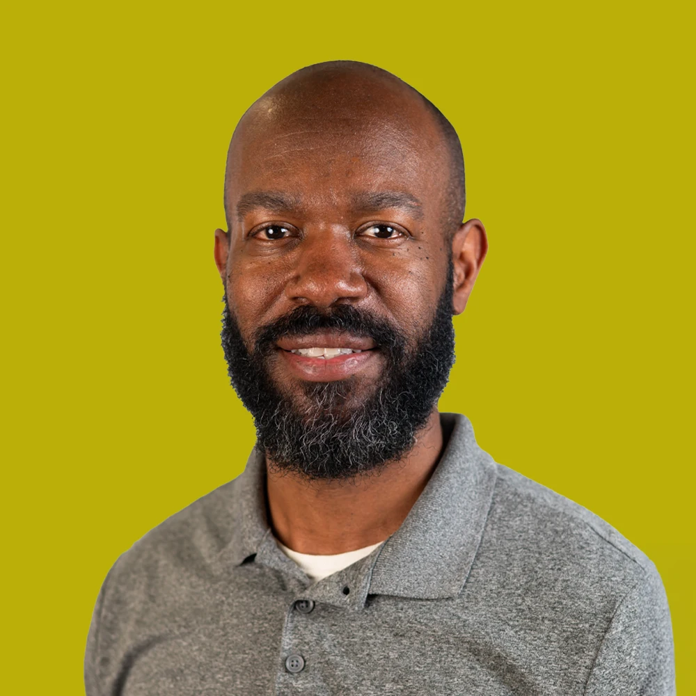 A man with a beard wearing a gray polo shirt smiles at the camera in front of a solid yellow-green background.
