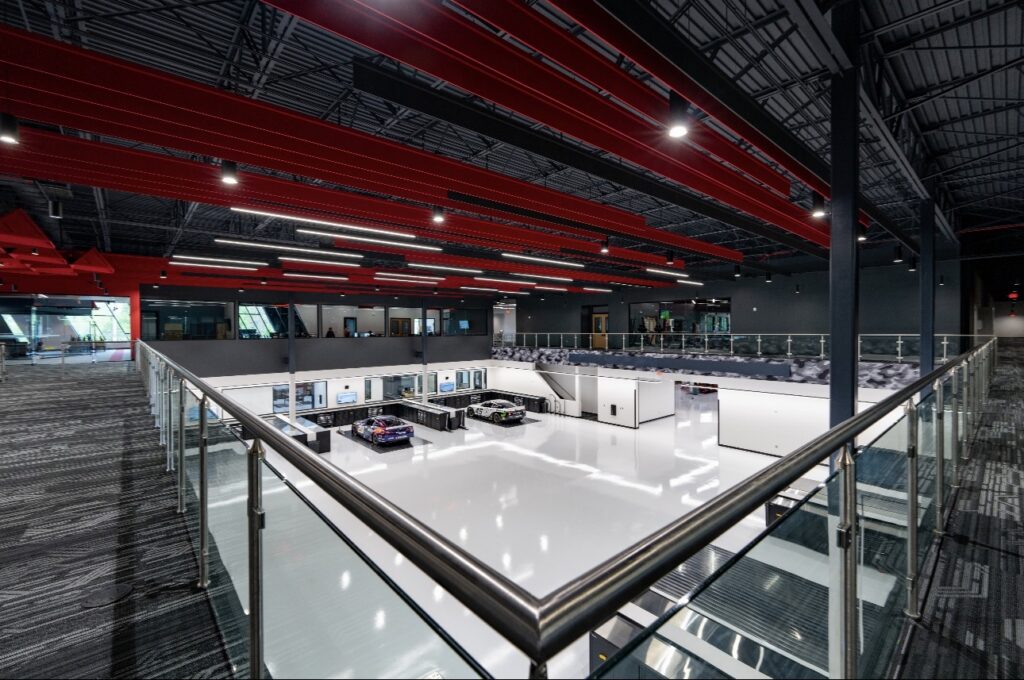 View of a modern industrial facility with white floors, large machinery, glass railings, and red ceiling beams under bright overhead lighting.